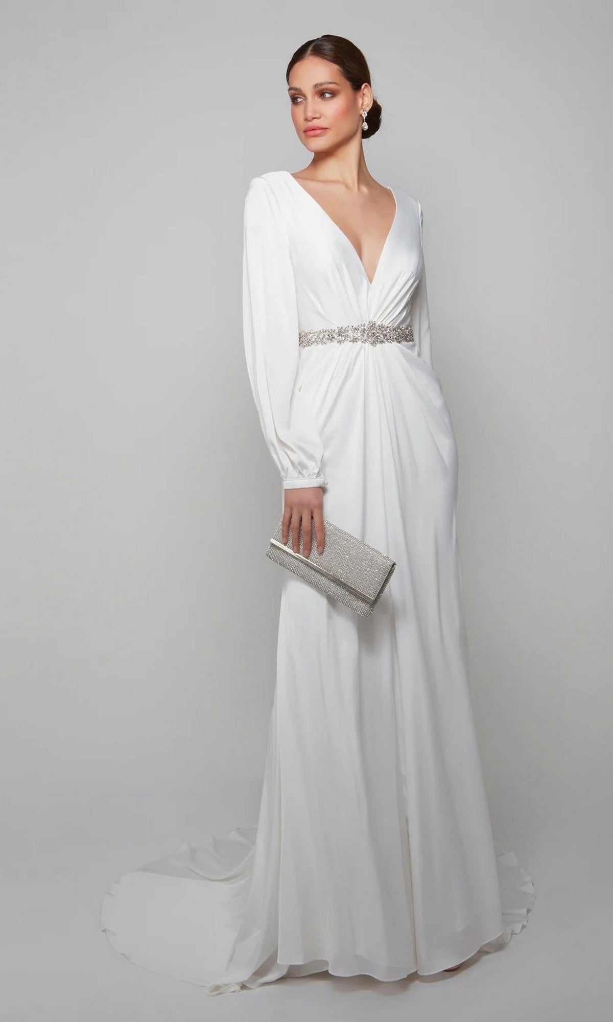 A woman models the Alyce 7070, a long-sleeved white satin formal gown with a deep V-neck and embellished waistband, holding a silver clutch against a plain light background.
